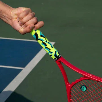 Red tennis racket with a yellow, green and black overgrip held by a person on a tennis court.
